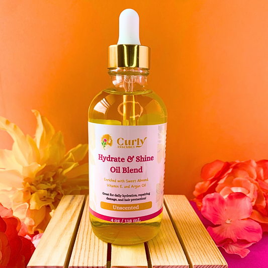 Front-facing bottle of Curly Assembly Unscented Hydrate & Shine Oil Blend posed with a floral background, offering a fragrance-free oil blend that deeply moisturizes, adds shine, and seals in hydration for healthy, shiny curly hair.