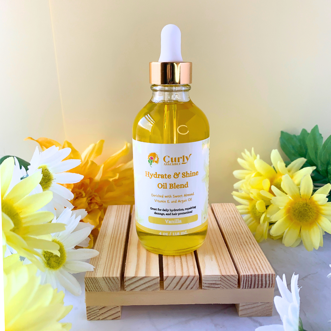 Front-facing bottle of Curly Assembly Sweet Vanilla Hydrate & Shine Oil Blend posed with a floral background, designed to deeply moisturize curly hair, add shine, and lock in hydration with a soft sweet vanilla scent.