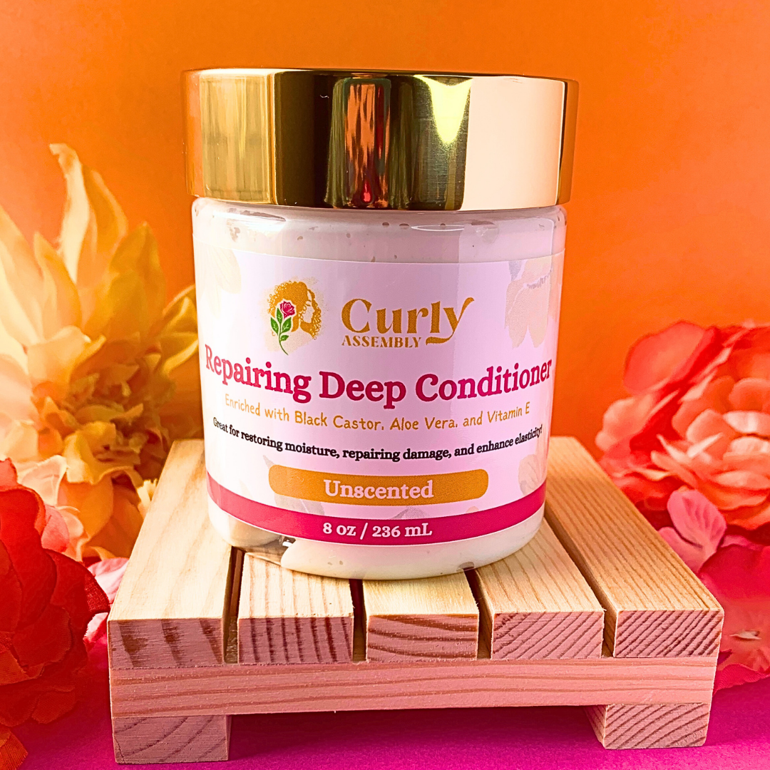 Front-facing jar of Curly Assembly Unscented Repairing Deep Conditioner posed with a floral background, offering a fragrance-free, deeply hydrating formula with aloe vera, black castor oil, and sweet almond oil to repair damaged curly hair and restore moisture.