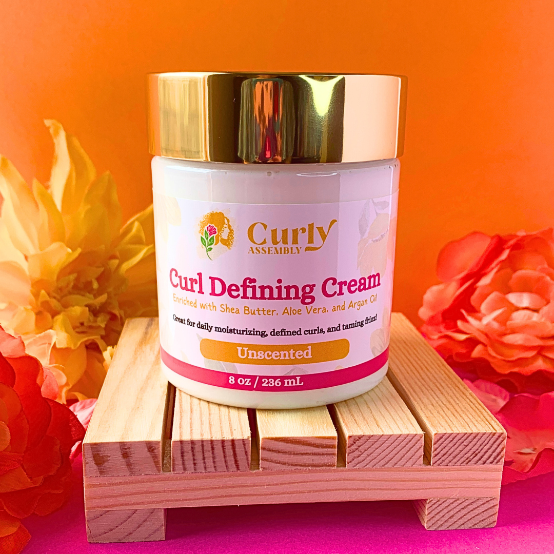 Front-facing jar of Curly Assembly Unscented Curl Defining Cream posed with a floral background, highlighting the fragrance-free option designed to nourish and define curls.